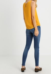 Mosterdgele mouwloze blouse met een sleutelgat aan de achterkant, gecombineerd met strakke blauwe jeans en platte schoenen met panterprint. Gladde stofstructuur.