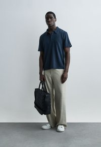 Άνδρας που φοράει μπλε ναυτικό πουλόβερ με κοντά μανίκια, μπεζ παντελόνια φαρδιάς γραμμής, λευκά αθλητικά παπούτσια, κρατώντας μια μαύρη τσάντα ώμου με ραφές, στέκεται μπροστά σε μια απλή τοιχο.