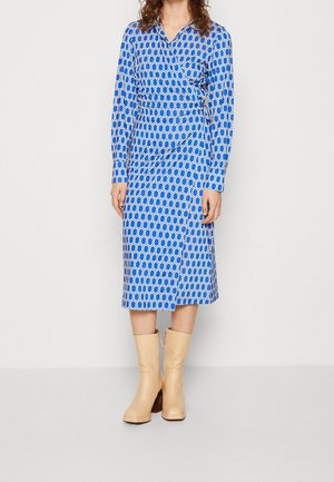 Blaues Kleid mit sechseckigem Muster, langen Ärmeln und einem Shirtkragen. Kombiniert mit beige Kniehochstiefeln mit einem kräftigen Absatz.
