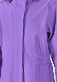 Veste violette présentant une texture lisse, une fermeture éclair, une taille élastique et un logo sur la poitrine. Conçue avec une capuche et des poches.