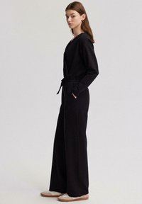 Mono negro con diseño envolvente, cintura elástica, mangas largas, bolsillos laterales y piernas anchas. Modelo viste zapatillas deportivas claras.