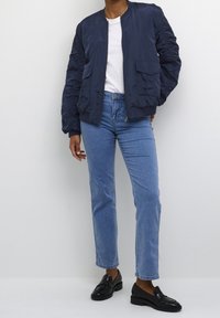 Marineblauwe bomberjack met glanzende afwerking, geribbelde kraag en twee voorzakken; gecombineerd met lichtblauwe jeans en zwarte instappers.