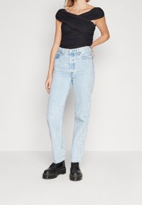 Černý top s krátkým rukávem a zkrouceným designem, kombinovaný se světle modrými džíny rovného střihu. Modelka má na sobě černé robustní boty.