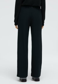 Schwarze, gerippte Hose mit weitem Bein, hoher Taille und strukturierter Stoff. Kombiniert mit schwarzem Schuhwerk, das die verlängerte Silhouette betont.