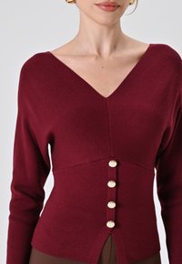 Maglione in maglia color borgogna con scollo a V, maniche lunghe e corpino aderente. Presenta cinque bottoni dorati sul davanti e fessure laterali.