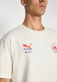 Krem majica od pamuka s crvenim Puma logotipom i plavim tekstom "KidSuper Studios", s grbom FC Salzburga sa strane. Okrugli izrez.