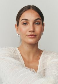 Sudraba aploces auskari, teksturēta balta topiņa ar sapilējušām detaļām, gluda āda un neitrāls grims gaišā fonā.