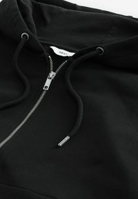 Sudadera negra con cremallera, capucha con cordón, cremallera metálica y tejido texturizado. Presenta una pequeña etiqueta de la marca dentro del cuello.