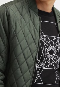 Veste bomber verte matelassée avec col côtelé et zip, superposée à une chemise noire présentant un motif géométrique blanc.