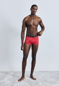 Piros boxer rövidnadrág fehér szívmintákkal, fekete rugalmas derékrésszel. A modell világosszürke felületen áll, minimalista háttérrel.