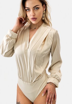 Femme aux longs cheveux blonds portant un body plissé beige et des boucles d'oreilles rectangulaires dorées, posant avec une main près de son visage et l'autre reposant sur la hanche.