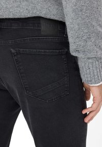 Jeans in denim nero con una texture liscia, dotati di una tasca posteriore cucita e un'etichetta in pelle sulla cintura.