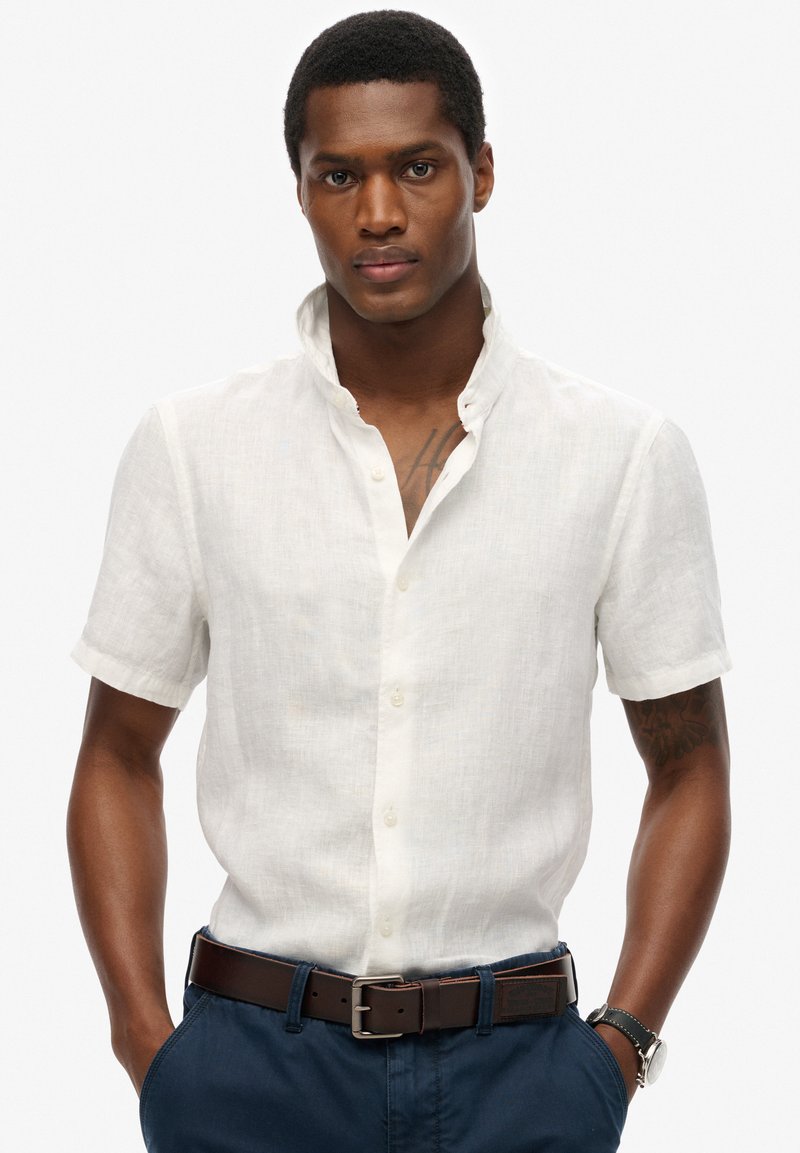 Hombre con cabello corto que lleva una camisa blanca de manga corta abotonada, pantalones azules, cinturón marrón, reloj de pulsera y un tatuaje visible en el pecho, con las manos en los bolsillos.