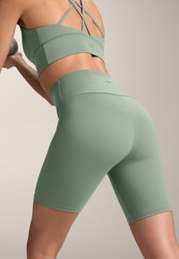 Olivovo zelený športový crop top a bicyklové šortky s vysokým pásom vyrobené z hladkej, elastickej tkaniny. Obsahujú dizajn krížových chrbátov a minimalistické logo.