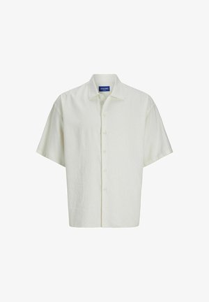 Short-sleeve, light beige button-up shirt with a relaxed fit, paired with navy shorts. Tan sneakers and white socks with text are worn.