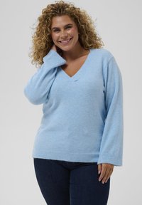 Pull bleu clair en tissu doux et texturé avec un col en V et des manches longues, associé à un jean bleu foncé.