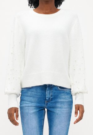 White sweater with textured finish and pearl accents on sleeves, featuring a round neckline and ribbed cuffs, paired with blue jeans.