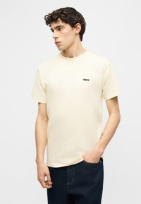 Νέος άνδρας με σγουρά μαλλιά που φοράει μια εκρού μπλούζα OBEY και σκούρο τζιν, κοιτάζοντας προς τα αριστερά με το ένα χέρι στην τσέπη του.