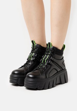 Bottes montantes noires avec lacets et accents vert néon, une semelle plateforme épaisse, et un mélange de matériaux synthétiques lisses et texturés.