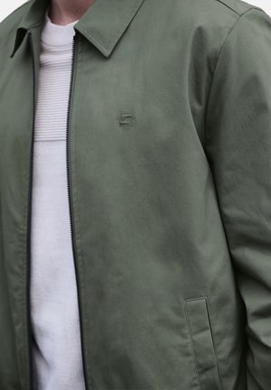 Hombre vistiendo chaqueta con cremallera color verde oliva sobre camisa blanca de canalé, con cuello visible y bolsillo frontal en la chaqueta.