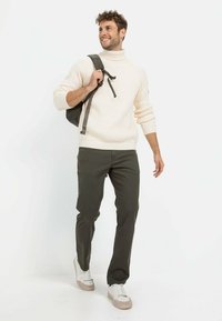 Cremfarbener, gerippter Rollkragenpullover, olivgrüne Hose und weiße Sneakers mit beigen Akzenten. Schwarzer Rucksack mit verstellbaren Trägern.