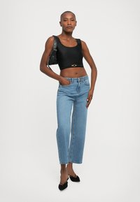 Woman wearing a black crop top, blue high-waist jeans, black slingback heels, and carrying a black shoulder bag against a white background.