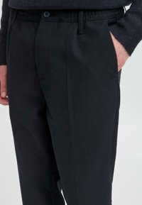 Schwarze maßgeschneiderte Hosen mit glatter Textur, seitlichen Taschen, elastischem Bund und Knopfverschluss, geeignet für formelle oder legere Anlässe.