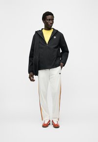 Άνδρας που φοράει μαύρο μπουφάν Adidas, κίτρινο πουκάμισο, λευκό παντελόνι με ρίγες στο πλάι και κόκκινα αθλητικά παπούτσια, στέκεται μπροστά σε λευκό απλό φόντο.