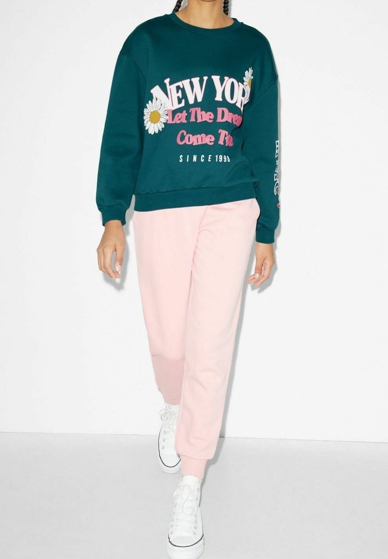 Personne portant un sweat-shirt bleu sarcelle avec le texte "New York Let The Dreams Come True" et des marguerites, un pantalon rose et des baskets blanches marchant vers l'avant.
