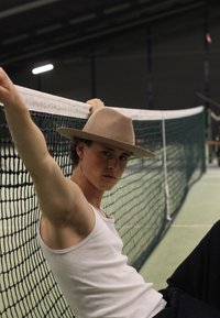 A young person leans against a tennis net, wearing a white tank top and a light brown felt hat, with dark pants and short hair.