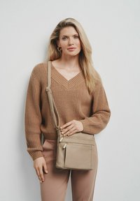 Beżowy sweter z dzianiny z dekoltem w szpic i długimi rękawami, zestawiony z jasnobeżowymi spodniami. W ręku trzymana jest mała beżowa torebka na ramię z zamkami.