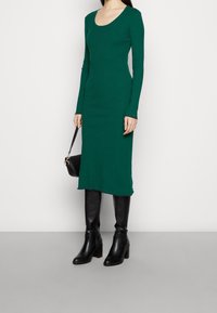Grön ribbad långärmad klänning med rundad halsringning, med en figursydd silhuett och rå fåll, kombinerad med knähöga svarta stövlar med klack.