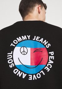 Homme portant une chemise noire avec un grand smiley coloré qui fait un clin d'œil et un symbole de paix, entouré des mots "Tommy Jeans Peace Love and Soul."