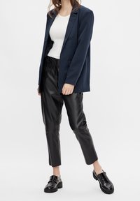 Blazer bleu marine, débardeur blanc côtelé, pantalon en cuir noir fuselé et chaussures noires massives. Le blazer est doté d'un col cranté et de poches.