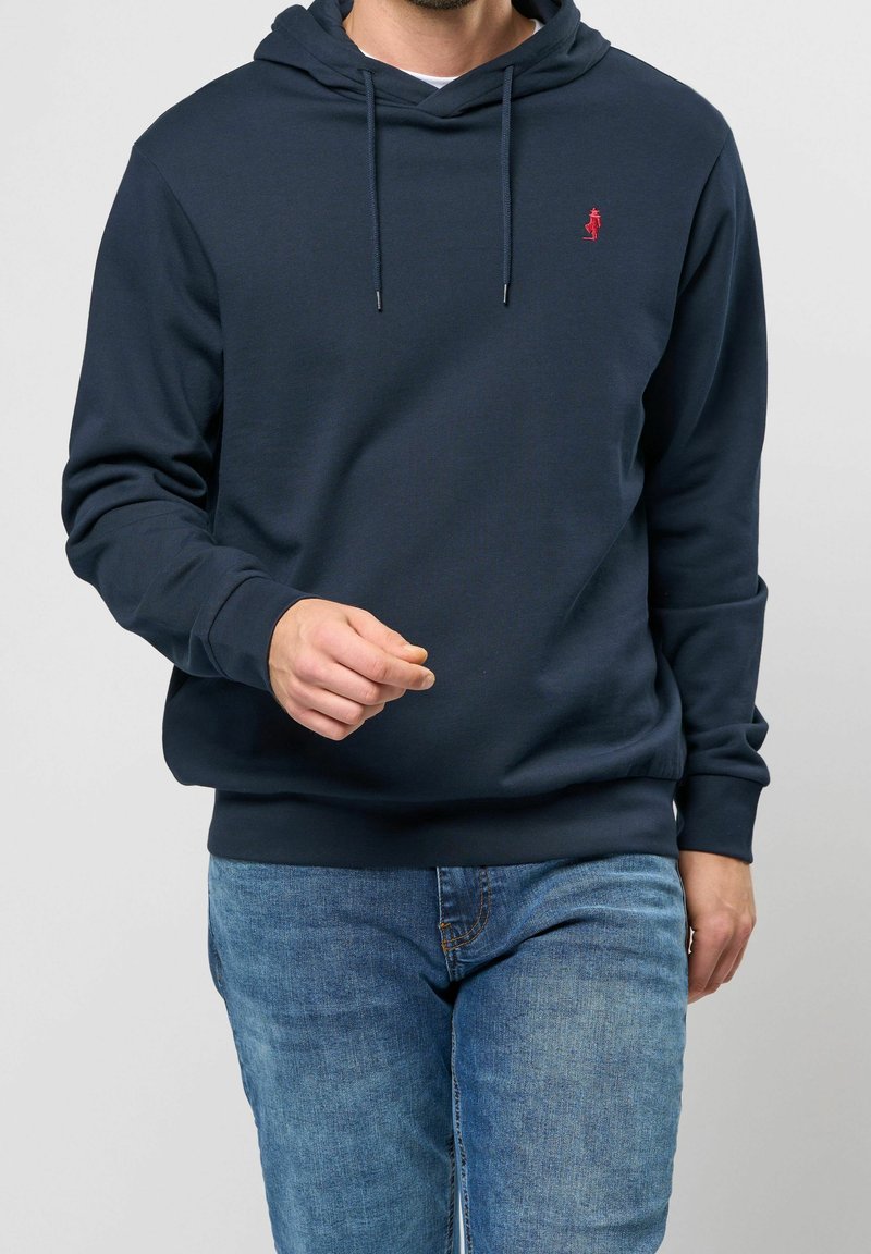 Sudadera azul marino con textura acanalada, que incluye una capucha con cordón. Pequeño logo rojo en el pecho, combinada con jeans de color azul claro.