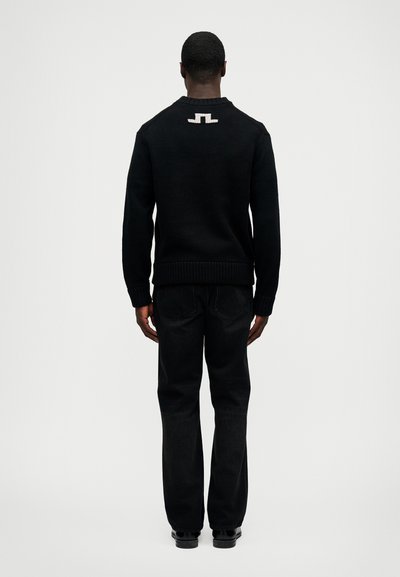 Fekete kötött pulóver bordás mandzsettákkal és aljjal, a hátán kontrasztos logóval. Sötét farmernadrággal és fekete cipőkkel párosítva.