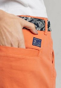 Pomarańczowe szorty bawełniane z wzorzystym czarnym paskiem w talii, z niebieską łatką logo na kieszeni i gładką fakturą.