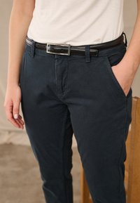 Persona, kas valkā baltu kreklu, iedurtu tumši zilos biksēs ar melnu ādas jostu, stāv ar vienu roku kabatā.