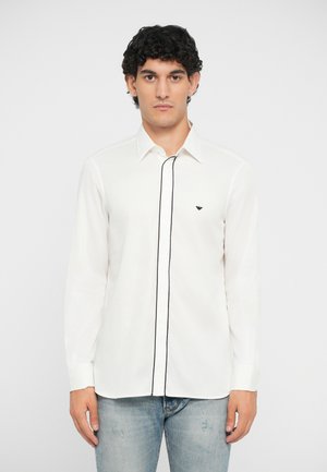 Camisa de manga larga blanca con ribetes negros a lo largo de la tapeta de botones, que presenta un pequeño logo negro en el pecho. La tela parece suave.