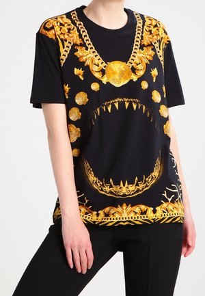 T-shirt noir avec chaînes baroques dorées et coquillages, avec un grand graphisme d'une bouche de requin ouverte au centre avant, porté avec un pantalon noir.