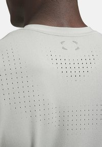 Camiseta deportiva gris claro con una textura suave, que presenta pequeñas perforaciones y un logo en la parte posterior del cuello.