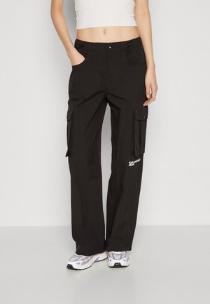 Person iført sorte cargobukser med for- og sidelommer, hvid cropped tanktop og hvide sneakers med lilla accenter.