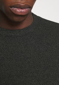 Pull texturé vert foncé avec un col rond côtelé. Gros plan montrant le motif de tricot complexe du tissu et sa finition lisse.