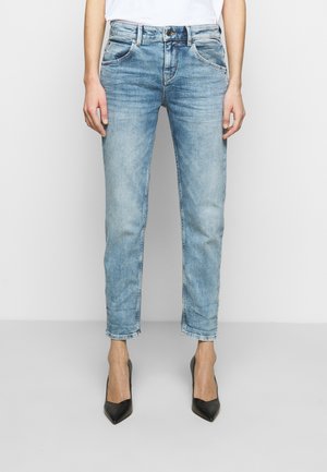 Vrouw die lichtblauwe cropped jeans en zwarte puntige hoge hakken draagt, staand tegen een effen lichte achtergrond.