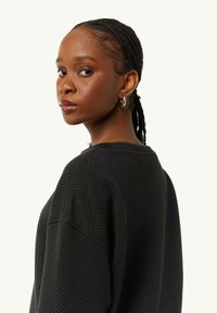 Schwarzer Pullover mit geometrischem Rippmuster, Rundhalsausschnitt und überschnittenen Schultern. Das Modell trägt silberne Creolen.