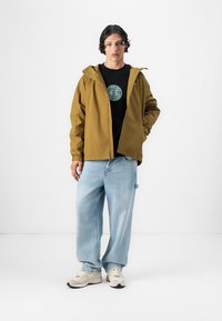 Khaki hættetrøje, sort t-shirt med cirkulært logo, lyseblå baggy jeans og beige sneakers. Glat stof og afslappet pasform.