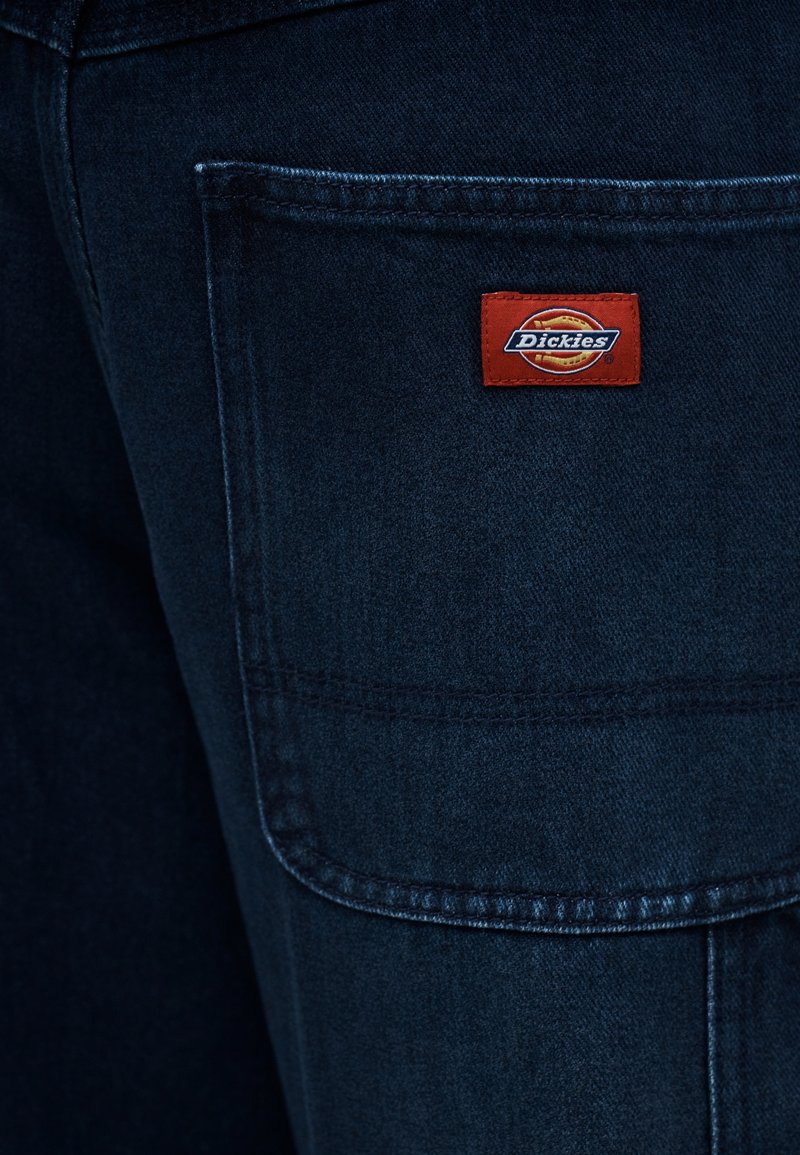 Poche de pantalon en denim bleu foncé avec un écusson rouge et jaune du logo de la marque Dickies cousu sur le rabat de la poche.