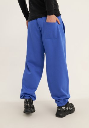 Person iført løstsiddende blå joggingbukser med elastiske manchetter og sorte sneakers, der står med den ene hånd i baglommen.