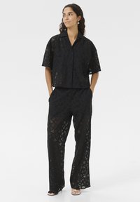Svart tvådelad outfit av genomskinlig spets med geometriska mönster; kortärmad skjorta med krage och löst sittande byxor.