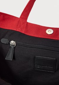 Czarna torba na ramię z czerwonymi materiałowymi uchwytami, wyposażona w zewnętrzną kieszeń na zamek, skórzane akcenty oraz ciemną metkę wewnątrz.